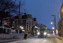 early morning with a brightening but dark sky on Duckworth Street show a heavy equipment snowblower blowing snow in a dump truck with a foreperson in a safety vest directing the snow removal operation